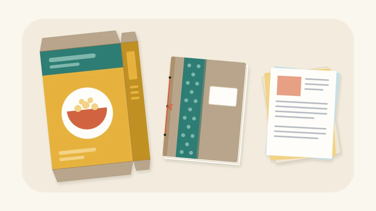 A small handmade notebook with a cardboard cover beside a flattened cereal box and a stack of scrap paper